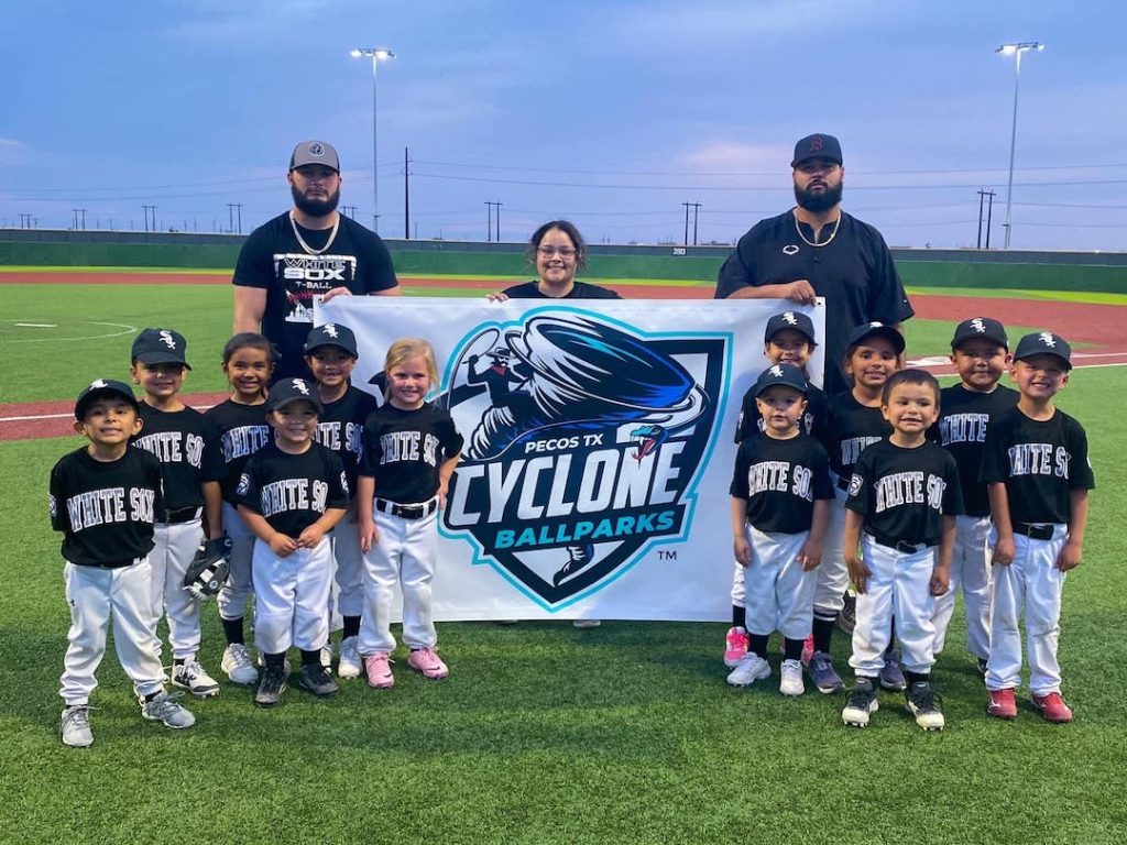 A team of youth baseball players and coaches from the Pecos Little League White Sox pose with a Cyclone Ballparks banner.
